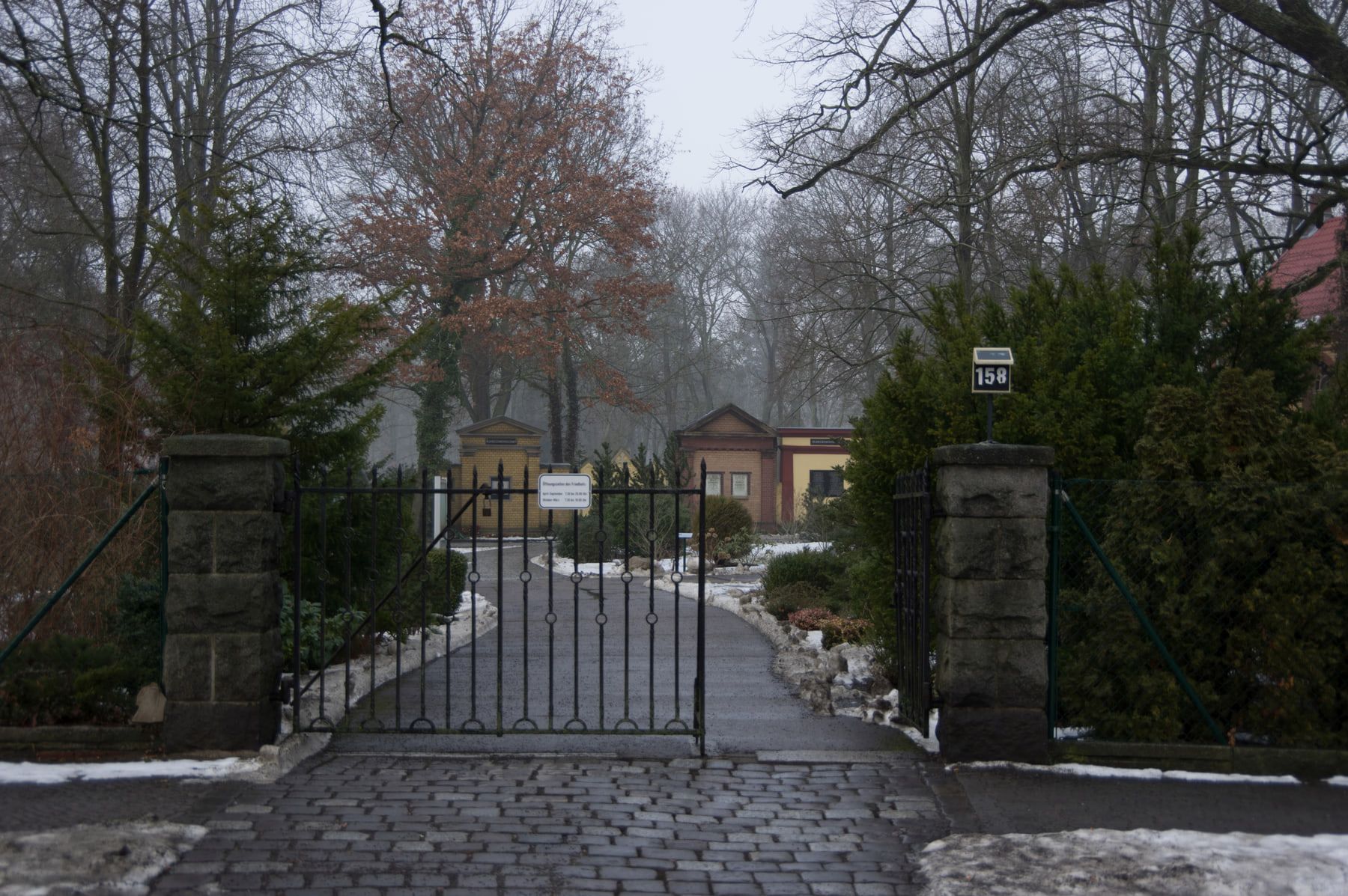 Wrought-iron entrance gate between stone pillars on a wintry path at Adlershof Cemetery