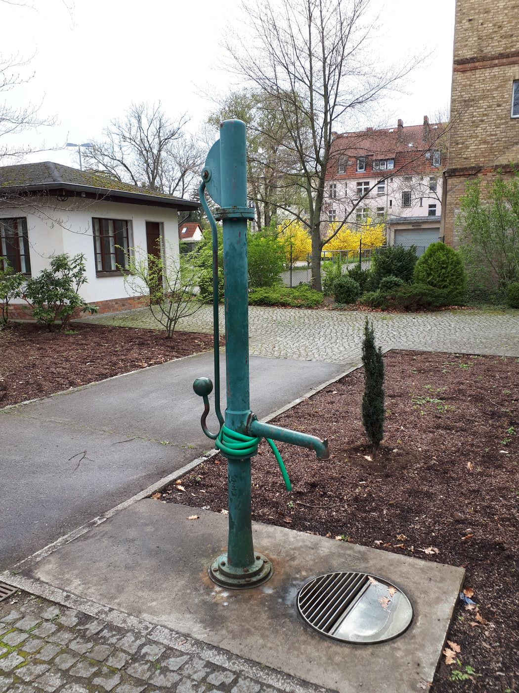 Green hand-operated water pump beside a paved cemetery path at Adlershof Cemetery